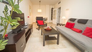 A living room with a grey sofa, red cushions, a television on a dark cabinet, a dining area with chairs, and a plant. - Marina de la Duquesa 722 (Manilva)