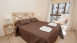 A neatly made bed with a brown bedspread and pillows, a wooden headboard, two bedside tables with lamps, and a chair by a window with a view of buildings. - Marina de la Duquesa 722 (Manilva)