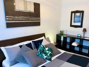 Habitación de hotel con cama, almohadas, mesita de noche, espejo y escritorio con lámpara.