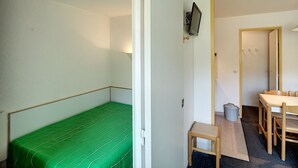 Una habitación pequeña con una cama verde, una mesa de madera con sillas y un televisor montado en la pared.