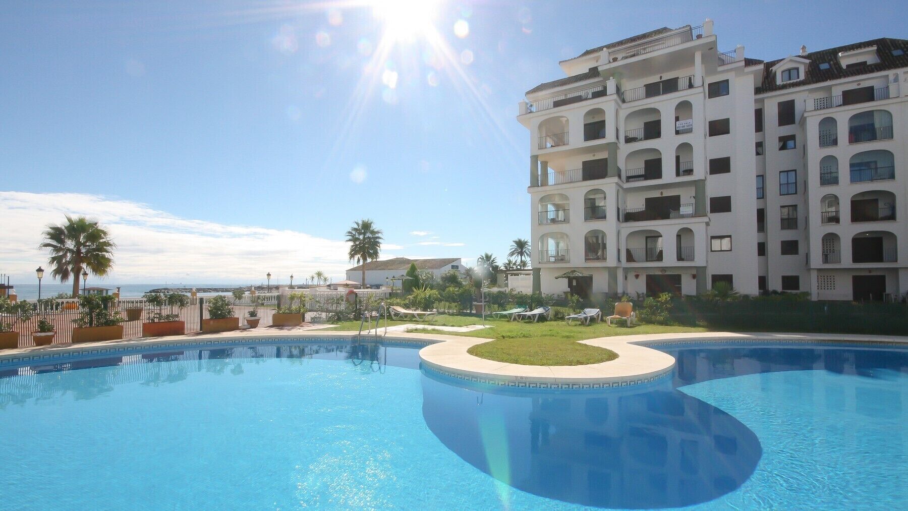 A swimming pool with a sunken center, surrounded by a white building with multiple balconies, palm trees, and a clear sky.