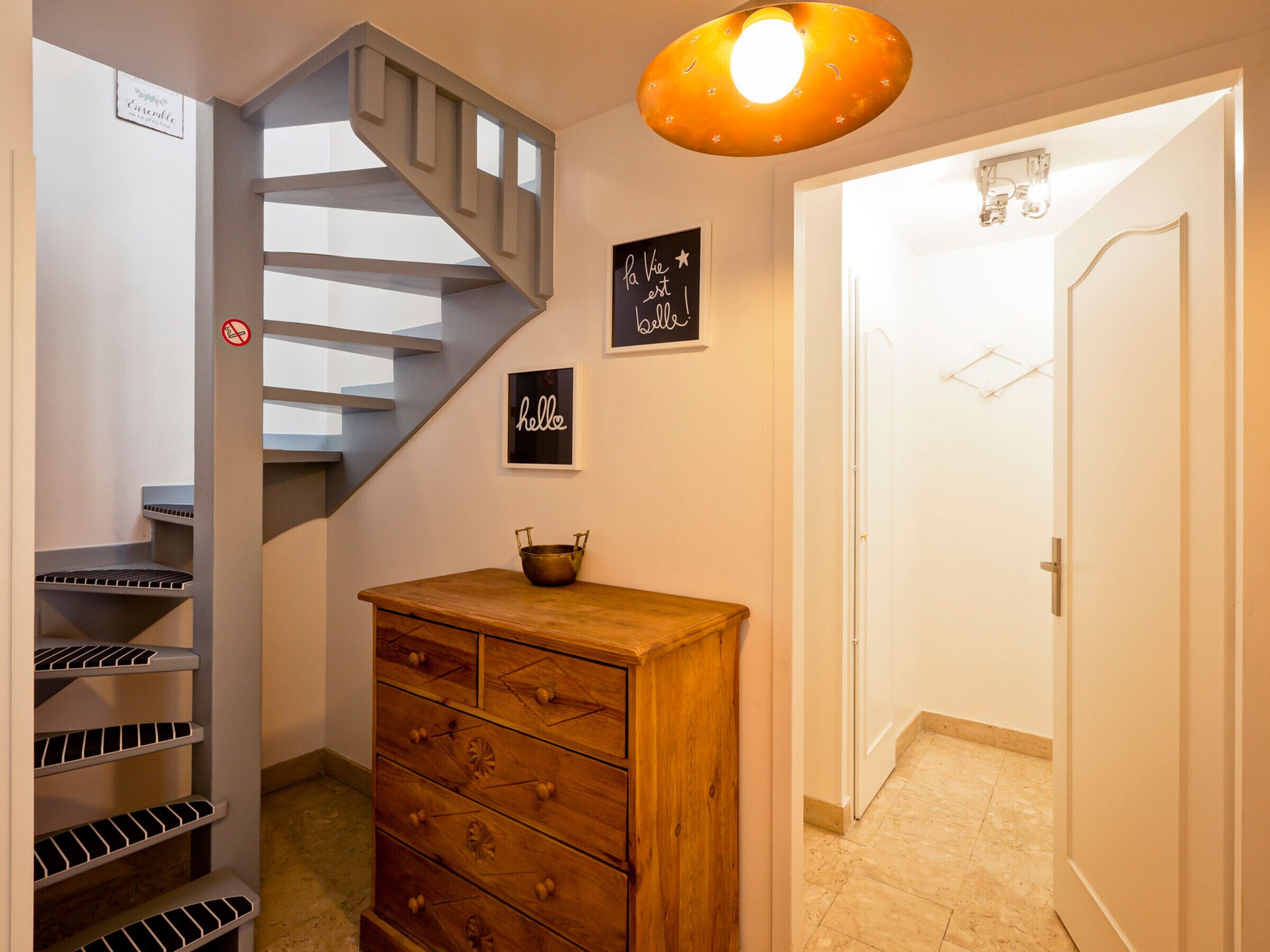 A staircase with a metal banister, a wooden dresser with drawers, a wall-mounted sign saying "hello", and a door leading to another room.