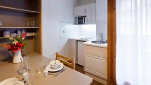 Une cuisine de taille réduite avec des meubles blancs, un four à micro-ondes et un évier. On y trouve une table à manger ornée d’un vase de fleurs et de quelques verres vides.