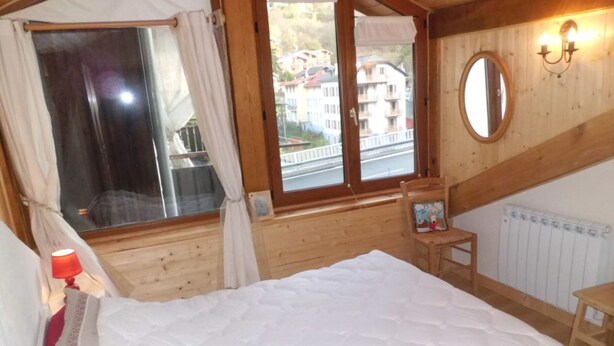 Quarto aconchegante com cama grande, janela com cortinas, cadeira de madeira, mesa pequena e espelho fixado na parede.