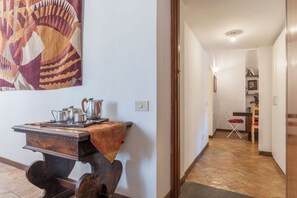 A hallway with a wooden table and metal teapots, a patterned tapestry on the wall, and a dining area in the background.