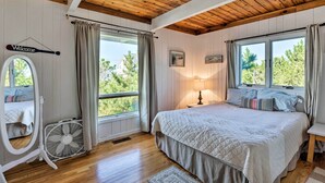 A bedroom with a wooden ceiling, a bed with a quilted bedspread, a bedside table with a lamp, a window with curtains, and a fan. - Westhampton Beach Home w/ Deck + Ocean Views! (Westhampton Beach)