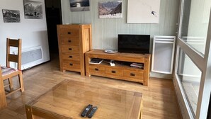 A living room with a wooden TV stand, a glass coffee table, and a chair. There are wall-mounted pictures and a window with a view.