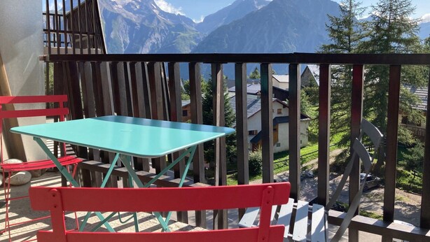 A balcony with a turquoise table and red chairs, overlooking a mountain range.