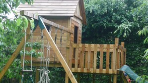 Ein hölzernes Spielhäuschen mit Rutsche und Kletternetz, umgeben von viel Grün.