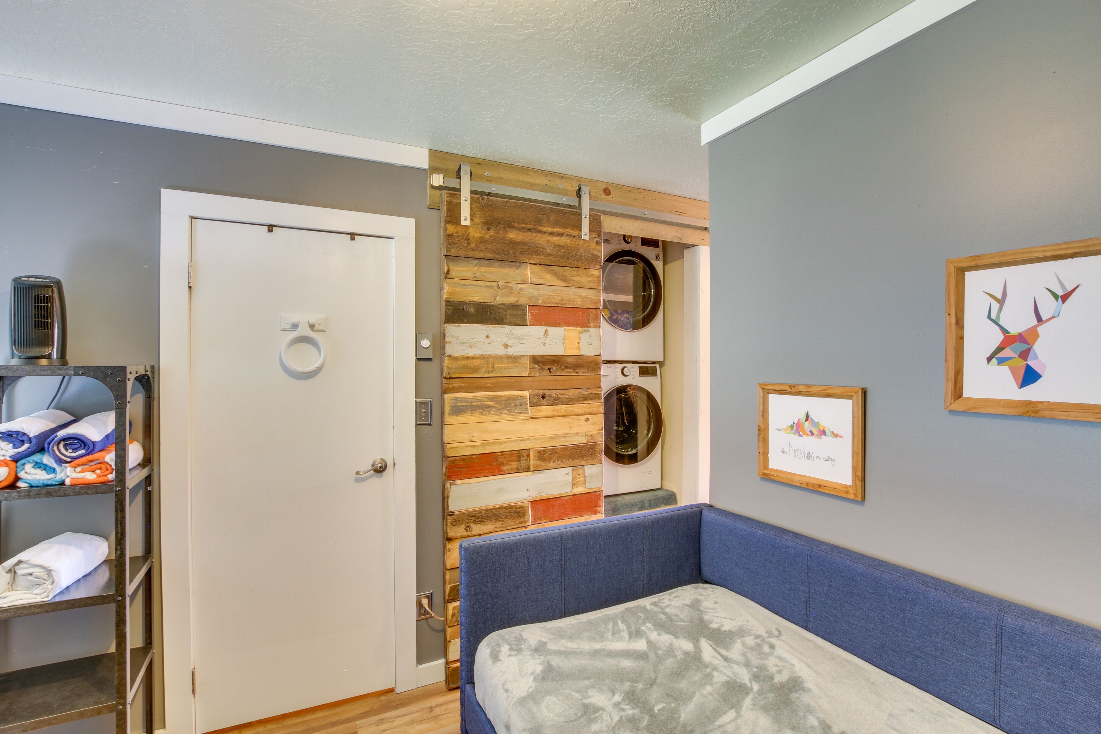 A room with a blue sofa, a wooden wall panel, a washing machine, a shelf with folded towels, and a framed picture on the wall.