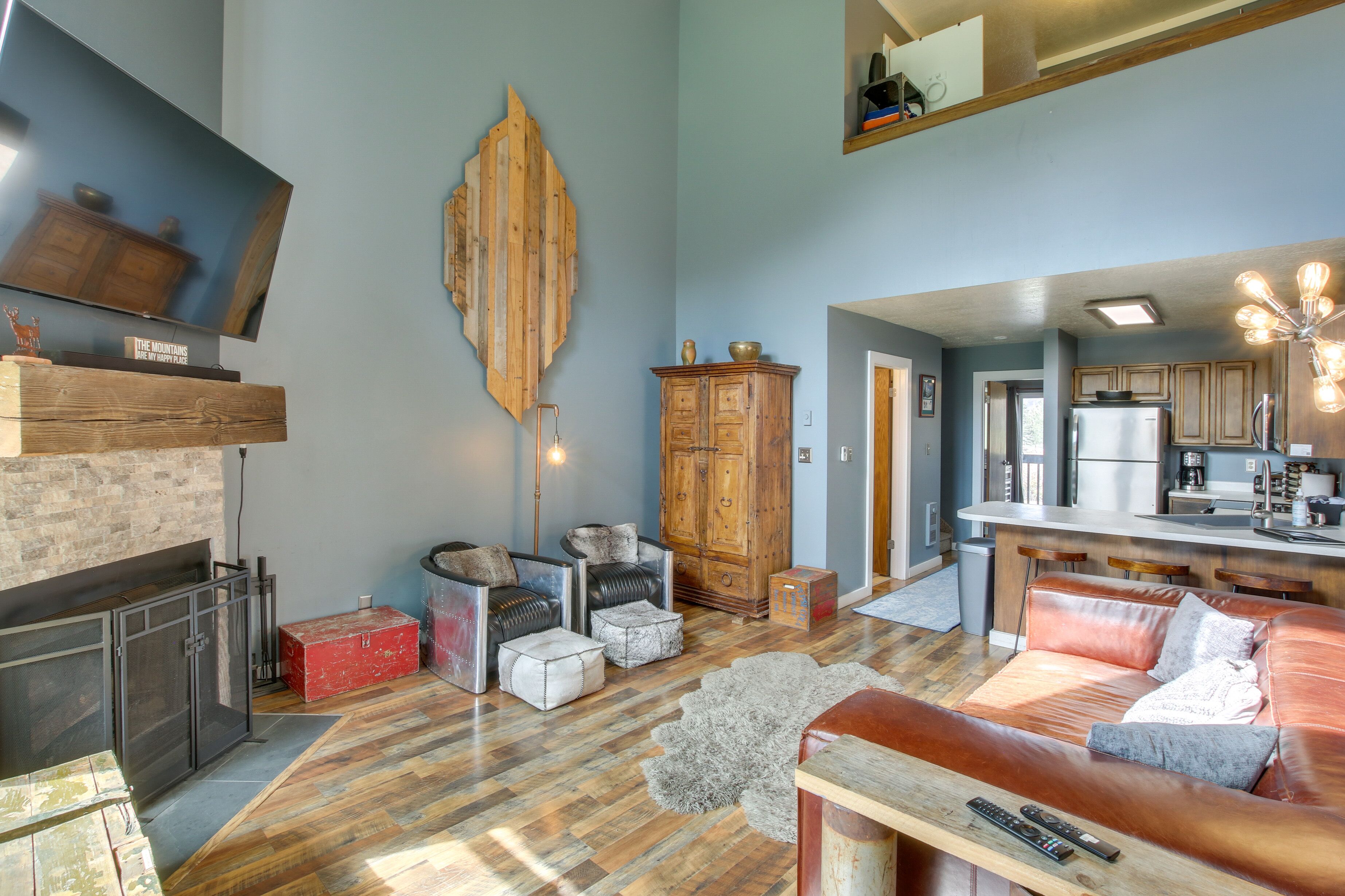 A modern living room with a fireplace, wooden cabinets, a large flat-screen TV, and a kitchen area with a refrigerator and bar stools.