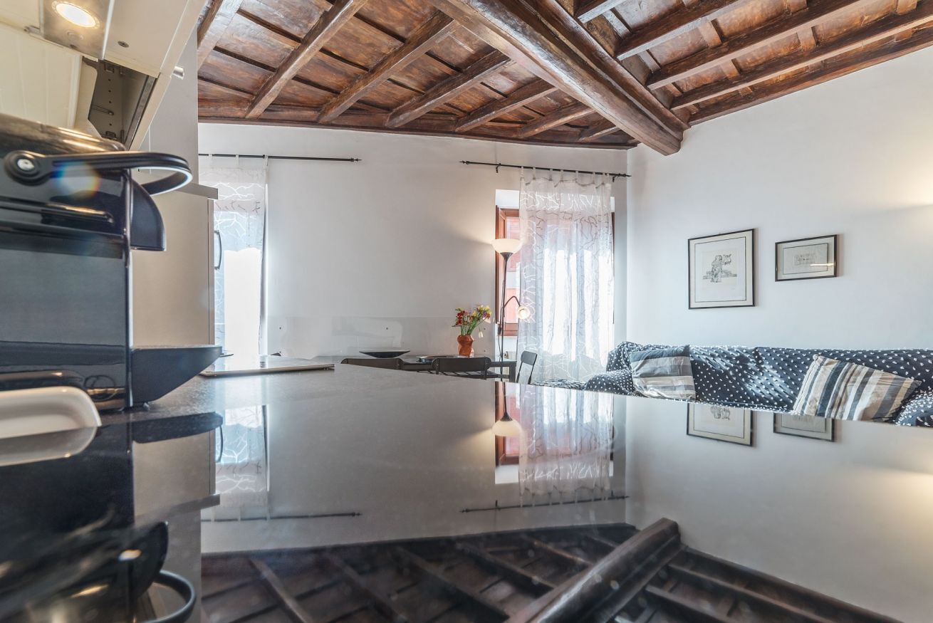A modern kitchen with a large island, wooden ceiling beams, and a dining area with a sofa and curtains.