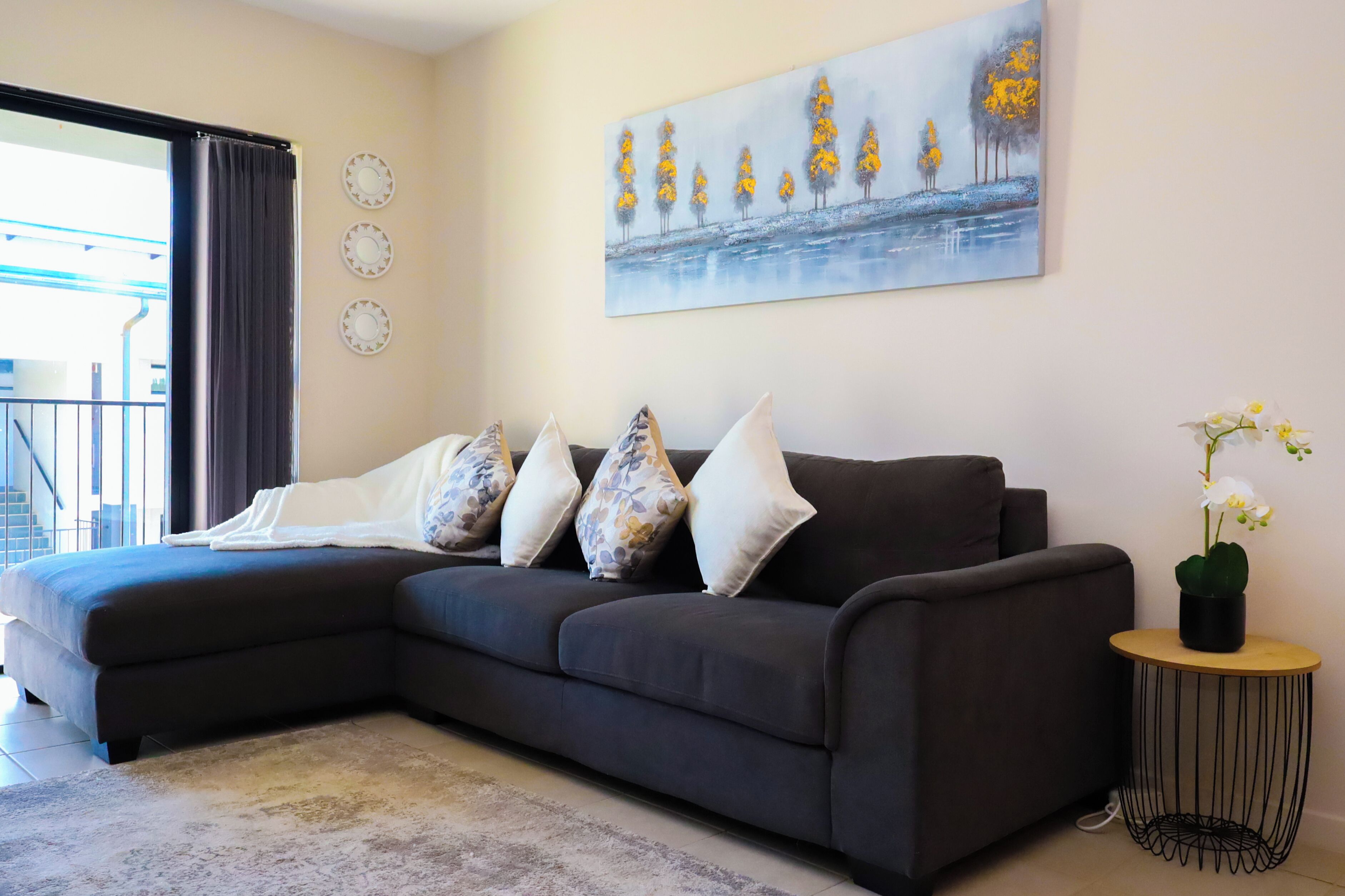 A modern living room with a dark grey sectional sofa, three decorative pillows, a round side table with a potted orchid, and a large landscape painting on the wall.