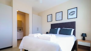 Una cama bien tendida con sábanas blancas, cabecera oscura y dos almohadas. Hay cuadros enmarcados en la pared, una mesita de noche con una lámpara y un pequeño armario.