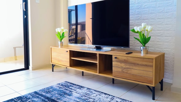 Una sala moderna con un televisor de pantalla plana sobre una consola de madera, dos plantas en macetas y un ventanal con vistas a un edificio.