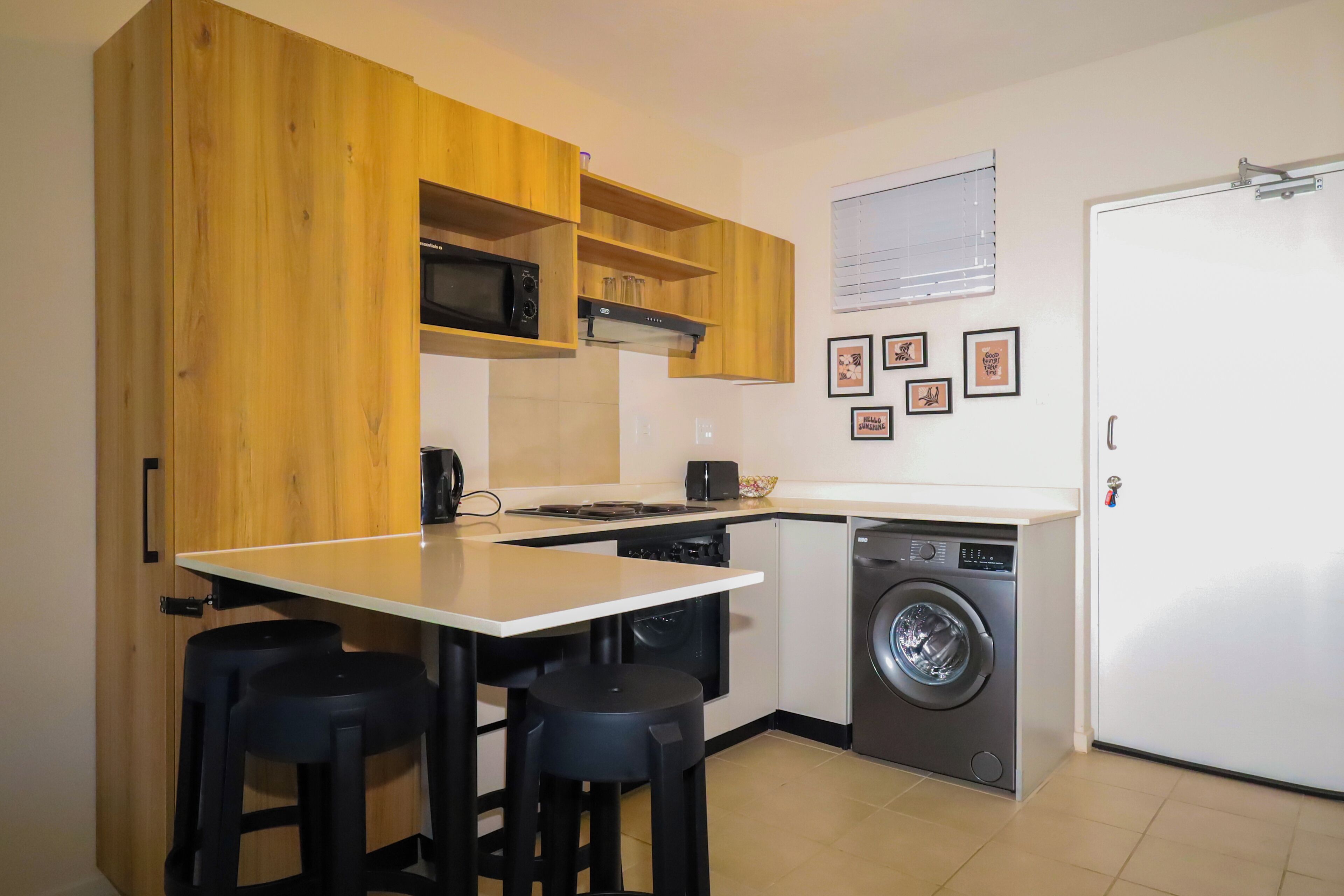 A compact kitchen with wooden cabinets, a microwave, a washing machine, and a small dining area with stools.