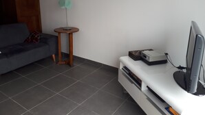 Uma sala de estar com um sofá cinzento, uma pequena mesa de apoio de madeira, um móvel branco para aparelhos eletrônicos e uma televisão.
