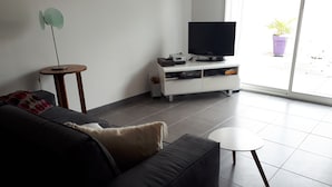 Uma sala de estar moderna com um sofá escuro, uma pequena mesa redonda, uma TV em um móvel branco e uma planta em vaso perto da janela.