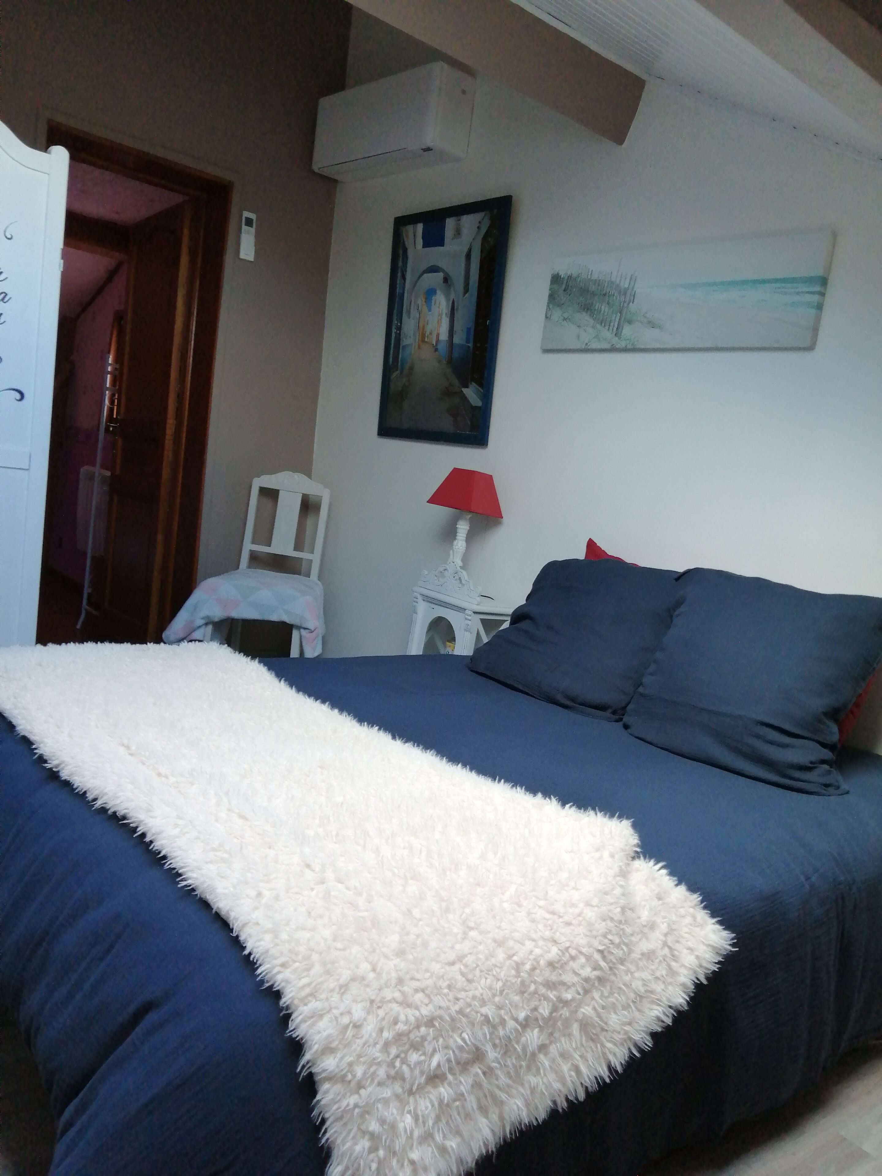 A bedroom with a bed, a white fluffy throw, a red lamp, and a painting on the wall.