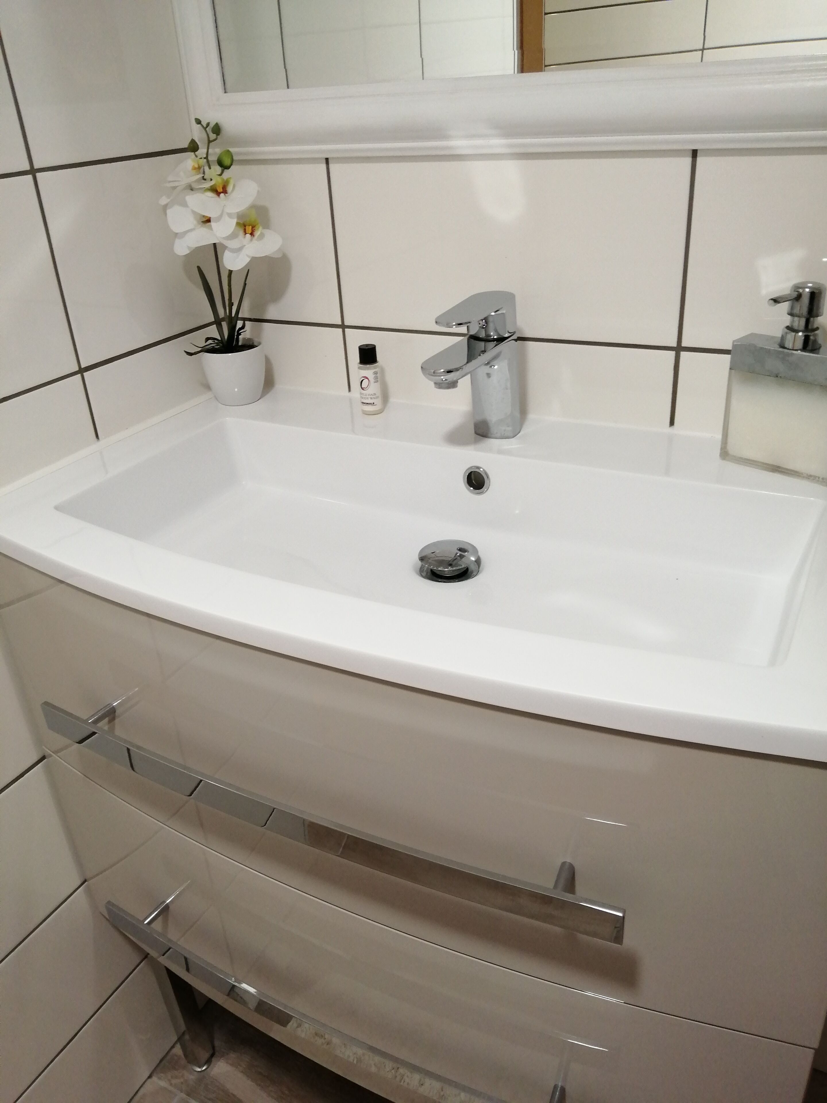 Un baño con un lavamanos blanco, un espejo encima, una orquídea en maceta y una botella de jabón para manos.