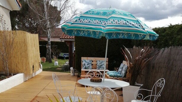 Une terrasse extérieure avec une plateforme en bois, des chaises en métal blanches, une table ronde et un grand parasol.