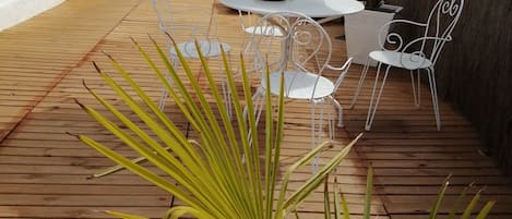 An outdoor patio with a wooden deck, white metal chairs, a round table, and a large umbrella.