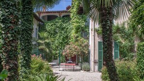 Un jardín con un sendero de grava que lleva a una mesa y sillas, rodeado de abundante vegetación y un edificio con contraventanas verdes.