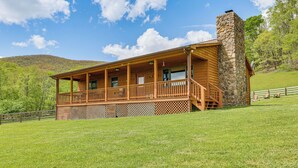 A wooden cabin with a stone chimney, a covered porch, and a view of a green hillside. - Fish, Hike, Hunt: Secluded Cabin in Doe Hill! (Doe Hill)