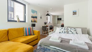 Una camera d'albergo con un divano giallo, un letto con cuscini bianchi e decorati, una scrivania con una sedia e vista sulla città attraverso una finestra.