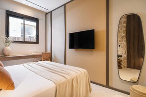 A modern bedroom with a bed, a flat-screen TV mounted on a textured wall, a large mirror, and a window with a view of a building.