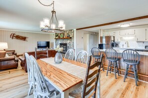 A spacious living room with a wooden dining table, white chairs, a brown leather sofa, and a TV.