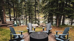 Un espace extérieur circulaire aménagé avec des chaises Adirondack disposées autour d’un foyer central, le tout entouré d’une zone boisée avec une terrasse en bois et des tables de pique-nique.