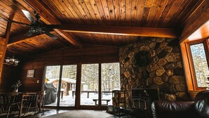 L’intérieur chaleureux d’un chalet avec des poutres apparentes, une cheminée en pierre et de grandes fenêtres offrant une vue sur le paysage extérieur.