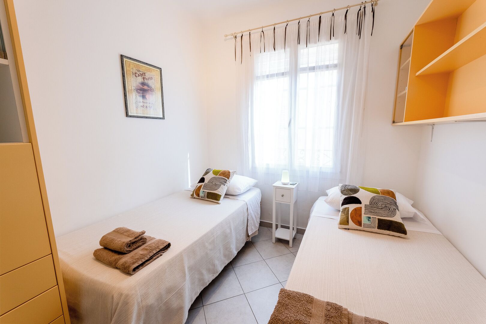 Habitación de hotel con dos camas individuales, una mesita de noche con lámpara, una ventana con cortinas ligeras y una fotografía en el marco en la pared.