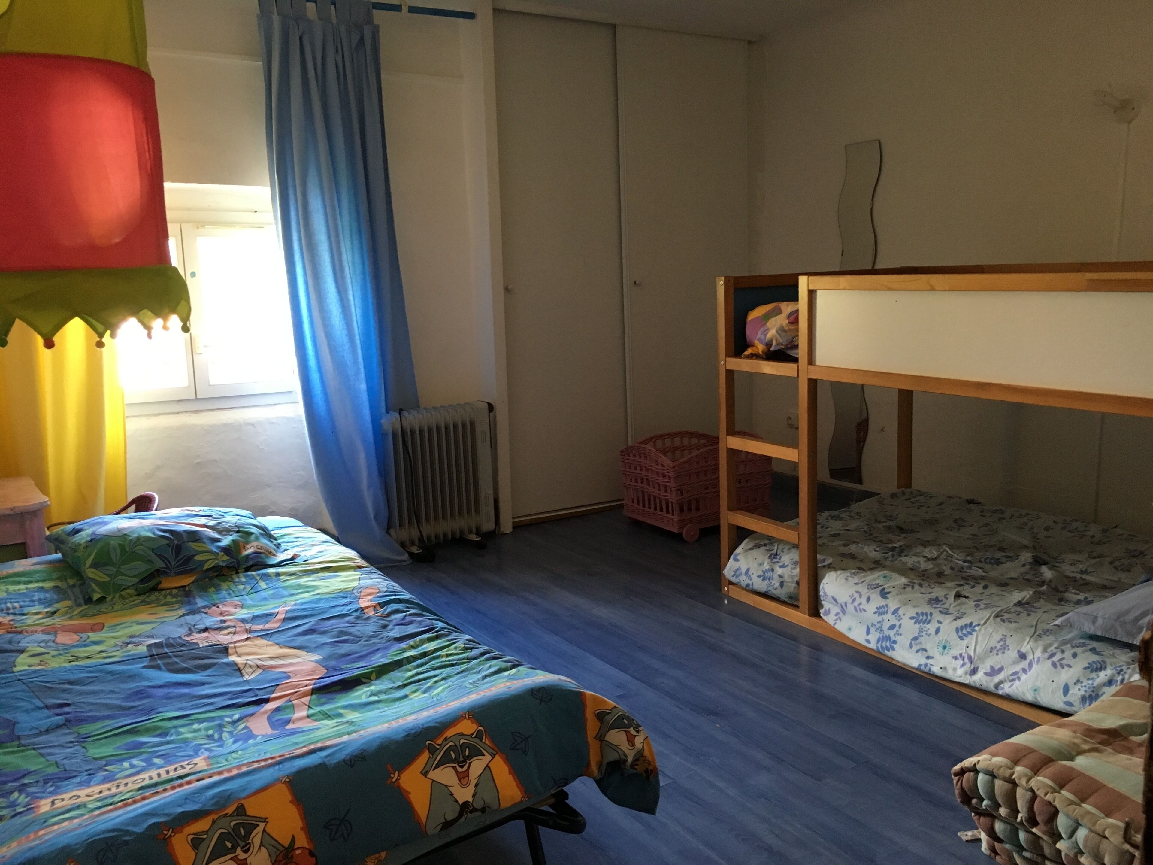 A room with a bunk bed, a single bed with a colorful bedspread, a radiator, a window with curtains, and a small stool.