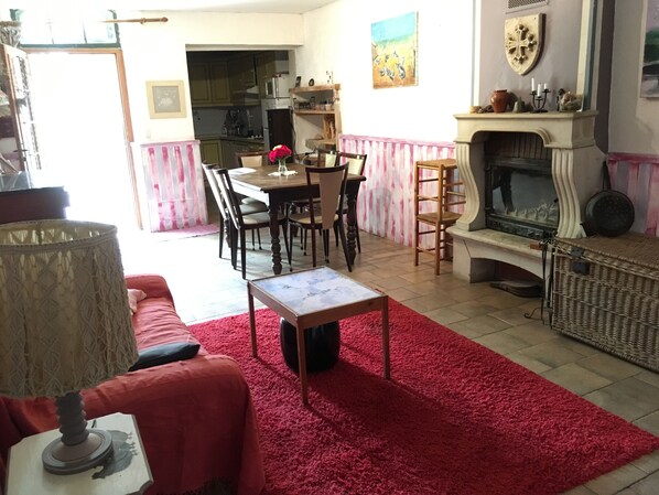 Een gezellige woonkamer met een rood tapijt, een open haard, een eethoek met een tafel en stoelen, en een keuken op de achtergrond.