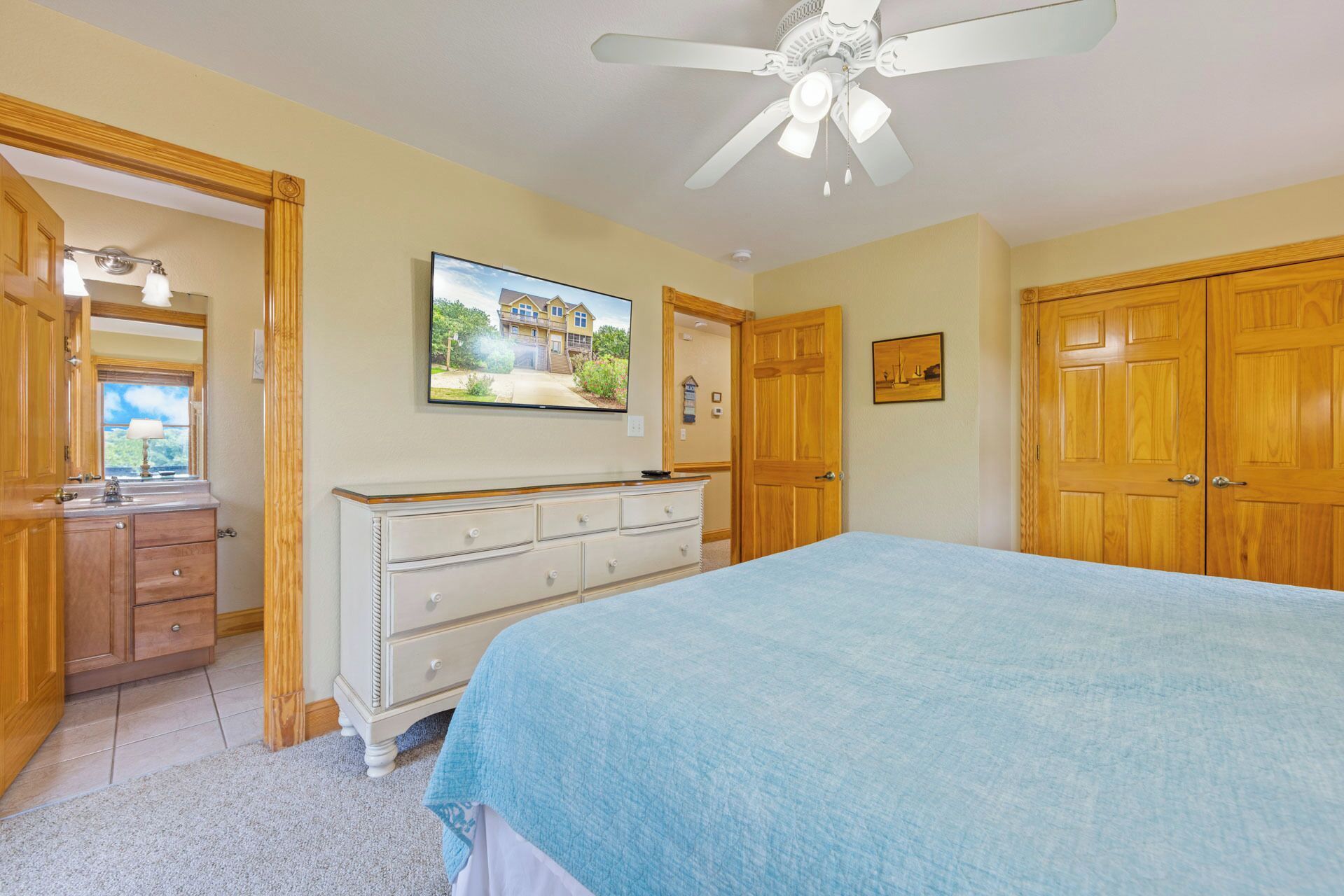 Une chambre à coucher comprenant un lit, une commode, une coiffeuse, un ventilateur de plafond et un téléviseur qui affiche une image de maison.