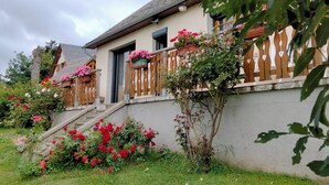 Une maison à deux étages avec une façade en pierre, des balustrades en bois sur le balcon et un petit jardin agrémenté de fleurs rouges.