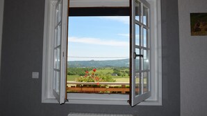 Une pièce dotée d’une grande fenêtre ouverte, offrant une vue imprenable sur un paysage verdoyant avec des collines et un ciel dégagé.