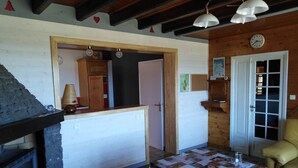Une pièce chaleureuse avec une cheminée en pierre, des poutres apparentes, une table recouverte d’une nappe matelassée et une horloge au mur.