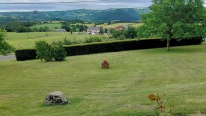 Un champ verdoyant avec un rocher, un parterre fleuri aux fleurs roses et, au loin, une vue sur des maisons et des collines.