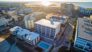 Vue aérienne d’une ville côtière avec différents bâtiments, dont un grand hôtel à plusieurs étages doté de balcons et d’une piscine.