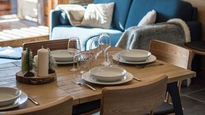 A dining area with a wooden table set for four, surrounded by chairs. A stone wall and a blue sofa are visible in the background. - Modern and warm apartment on the slopes, up to 6 guests (LA TOUSSUIRE)