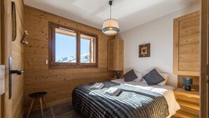A cozy bedroom with a wooden interior, a large bed with a striped blanket, two pillows, a bedside table with a lamp, and a window with a mountain view. - Modern and warm apartment on the slopes, up to 6 guests (LA TOUSSUIRE)