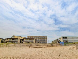 A beachfront hotel with multiple floors, a sandy beach in the foreground, and a clear sky.