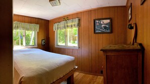 A room with wooden walls, a bed with a white bedspread, two windows with curtains, a painting on the wall, and a small wooden table. - Rangeley Waterfront Retreat: 4 Bed, 2 Bath, Perfect for Families to Unwind (Rangeley)