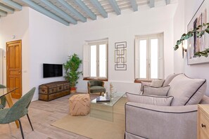 A spacious living room with a sofa, armchair, and a glass coffee table. There is a wooden cabinet and a television on the wall.