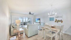 A modern living room with a sofa, dining table, and chairs. There is a ceiling fan, a TV, and a window with blinds. - Captiva Beach Villas 🏝 On The Gulf Beach (Captiva)