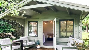 A cozy porch with two green chairs and a small table, a wooden deck, and a view of a garden.