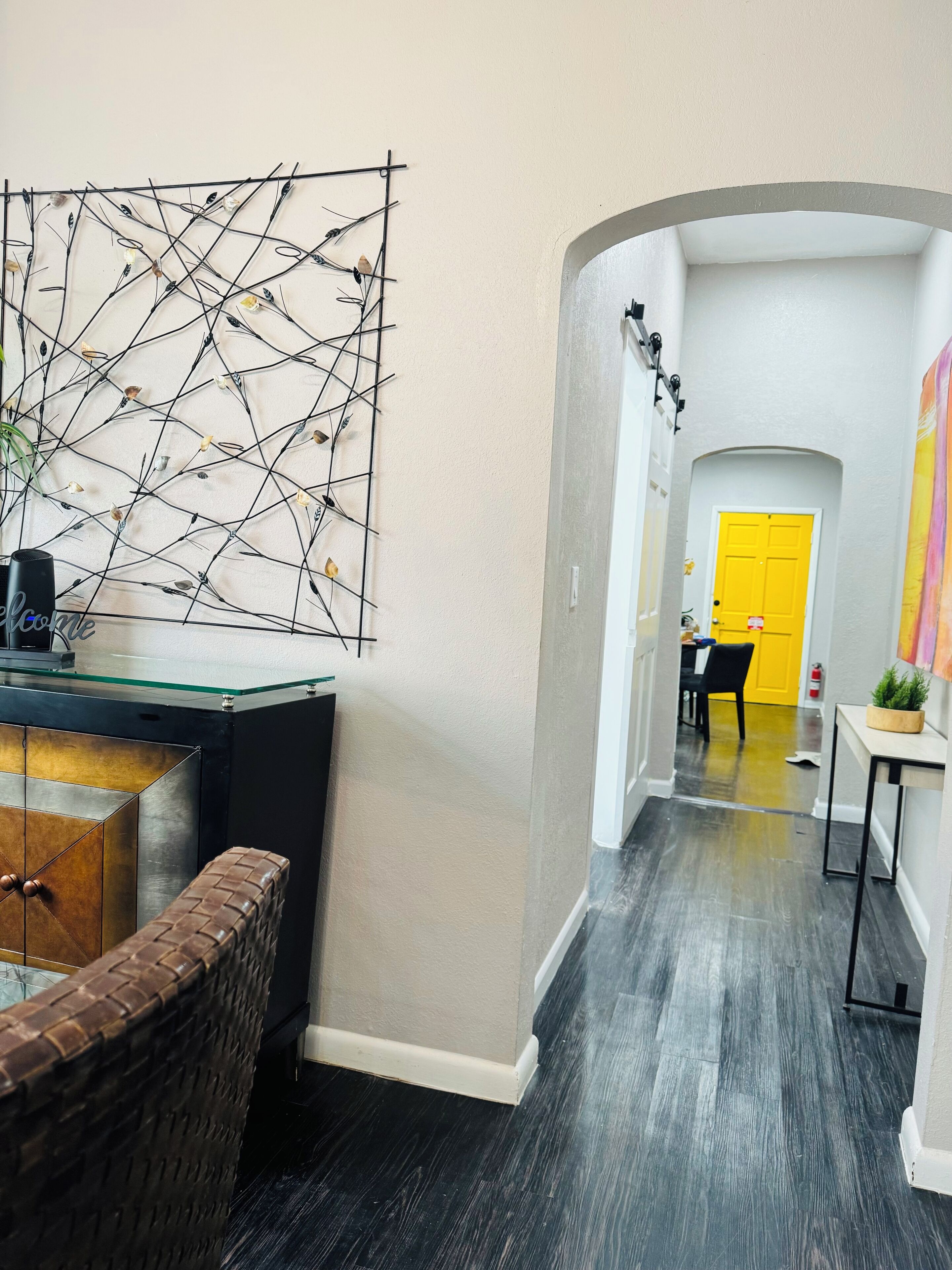 Um corredor com decoração moderna, incluindo uma obra de arte de metal preto na parede, uma mesa de centro de vidro e uma cadeira de vime.